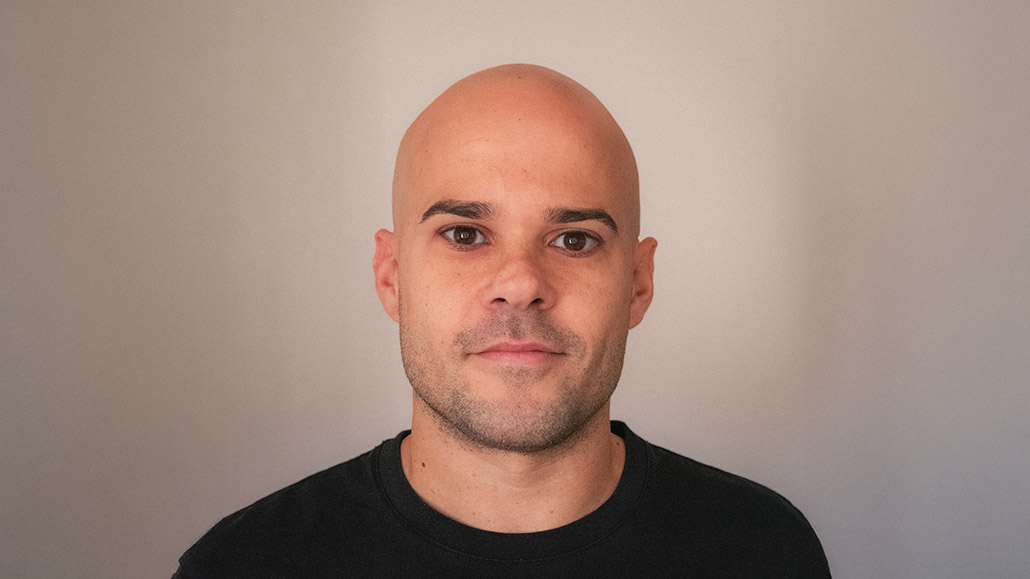 A man with a bald head and short beard, wearing a black t-shirt, stands against a light grey background.